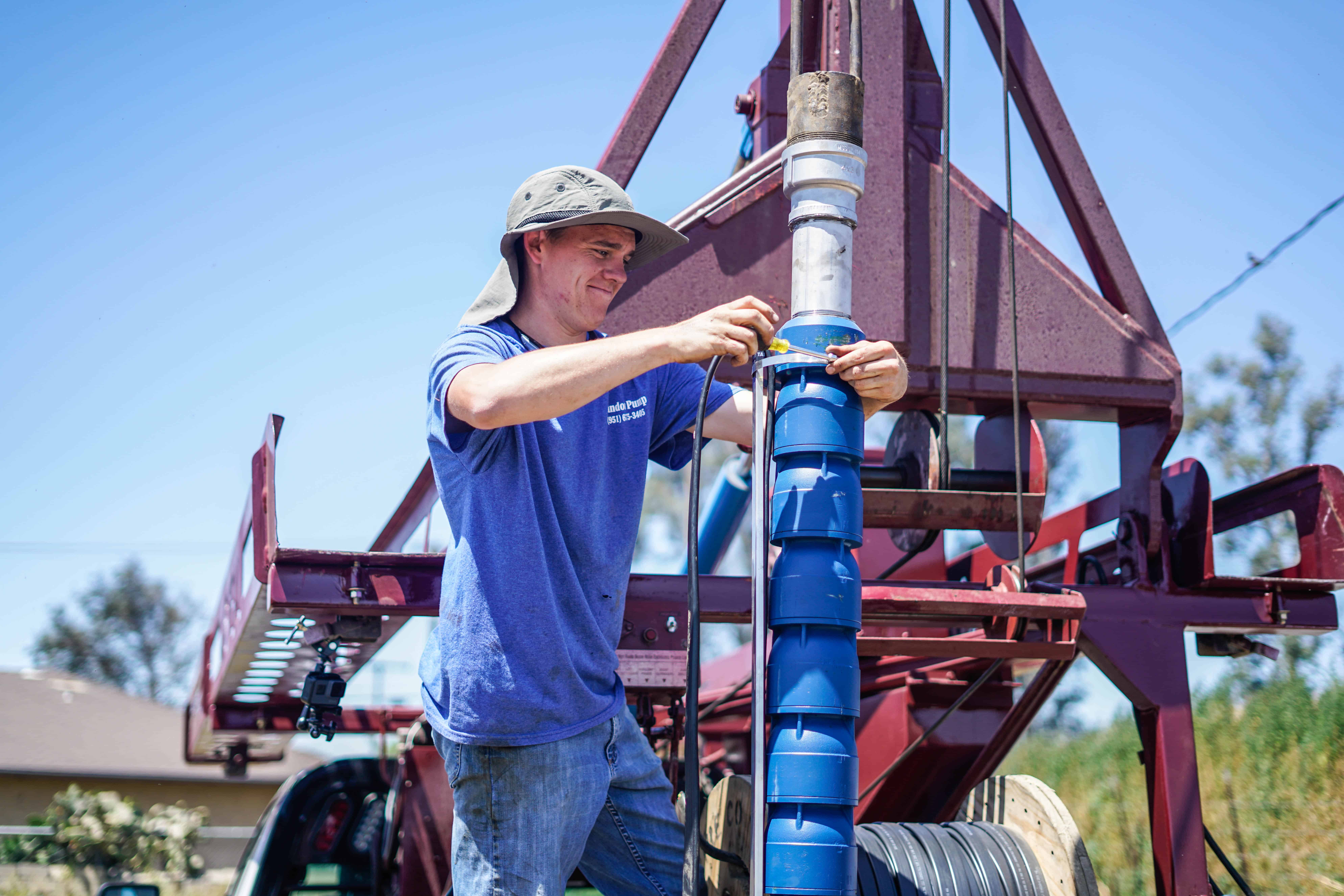 Technician in a blue shirt and hat installing a blue well pump on a drilling rig, ensuring proper water system setup.
