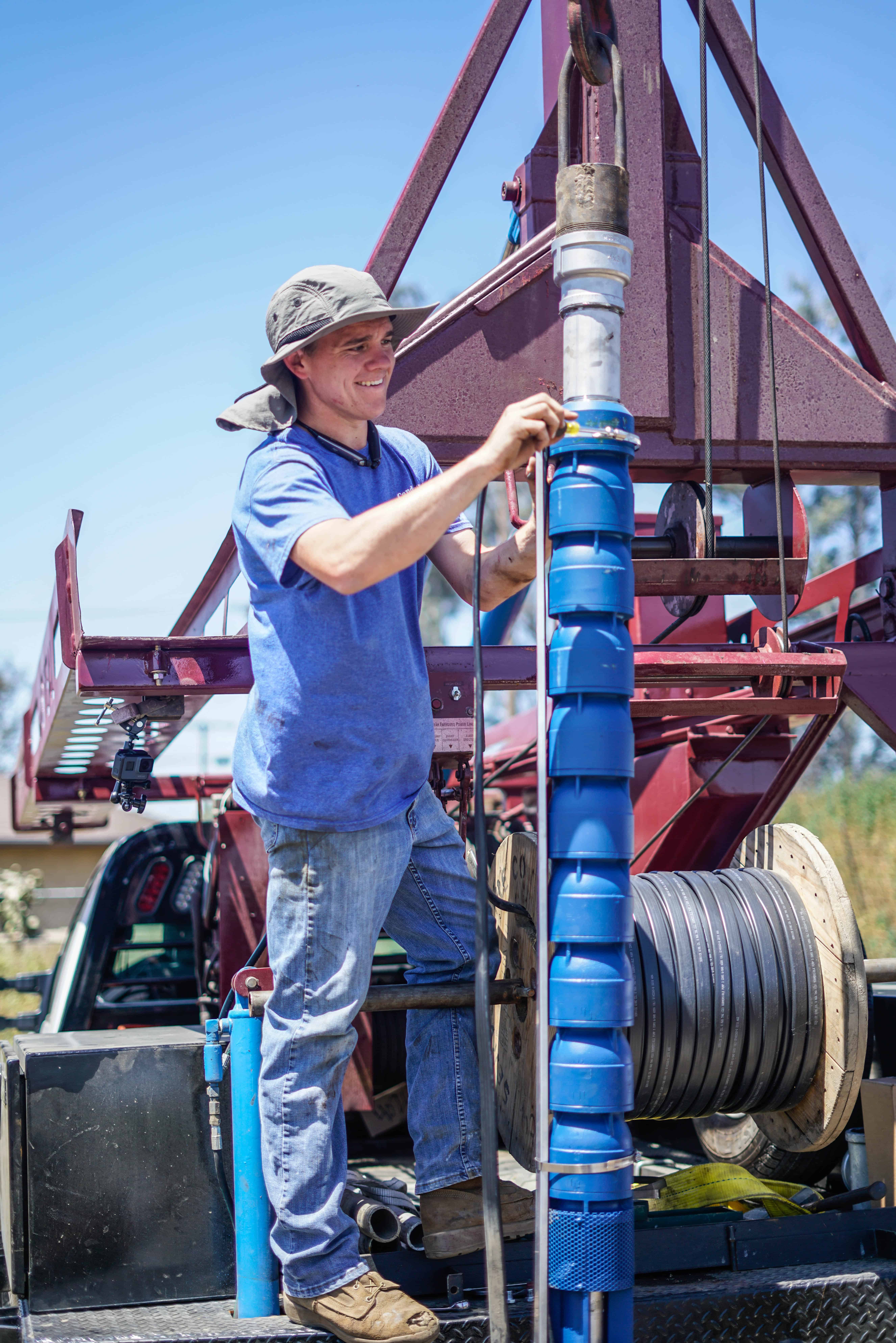 Technician in a blue shirt and hat installing a blue well pump on a drilling rig, ensuring proper water system setup.