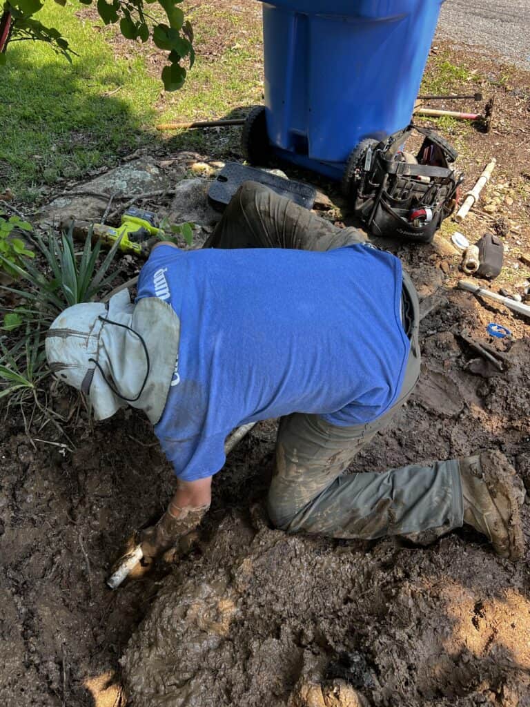 Technician in a blue shirt and hat repairing a buried water line in muddy conditions, using tools to access the underground piping.